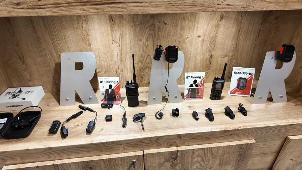 RayTalk Bluetooth wireless accessories displayed on a wooden table, including a Bluetooth speaker mic, Bluetooth PTT, Bluetooth transmitter and receiver, bone headset, and consumer headphones.