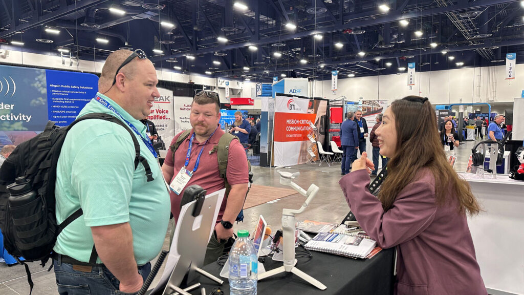 Two male visitors consulting a female salesperson at the booth about two-way radio earpieces and headsets